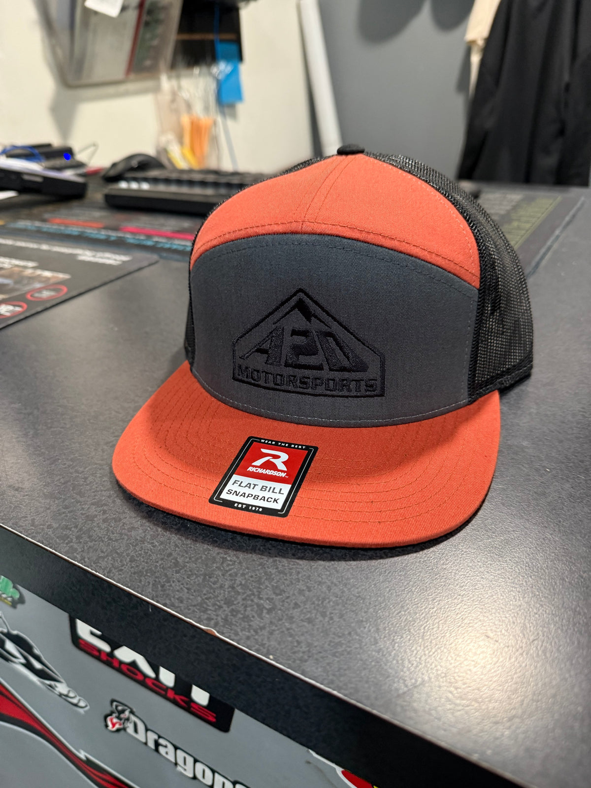 A2D Motorsports trucker hat, orange and charcoal flat bill snapback with black embroidered mountain logo